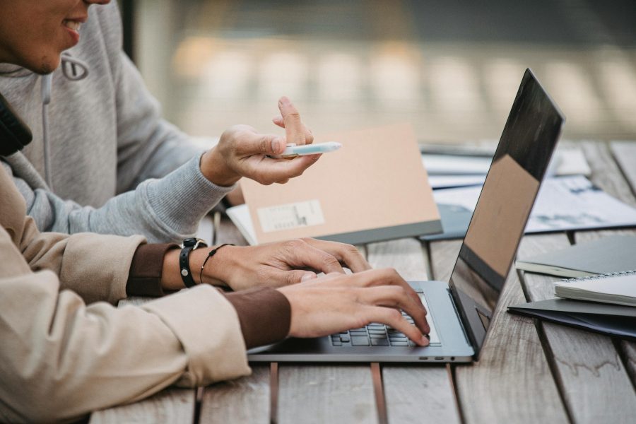 Person typing on laptop computer with another person sitting next to them holding a pen