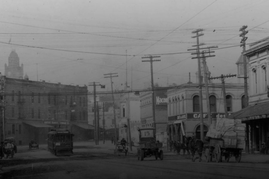 An image of the French district in downtown Los Angeles. The image shows Aliso Street in downtown Los Angeles, California, with signs labeling buildings "Griffins Transfer and Storage Co." and "Cafe des Alpes" next to "Eden Hotel," which are located on opposite corners of Aliso and Alameda Streets. A Pacific Electric streetcar sign reads "Sierra Madre" and automobiles and horse-drawn wagons are seen in the dirt road.