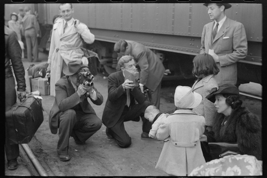 A black-and-white photo depicts cameramen snapping photos of a young Japanese girl as she waits to be sent from Los Angeles to the Manzanar prison camp.