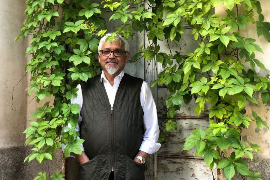 Amitav Ghosh stands amid vines against a white stone wall.