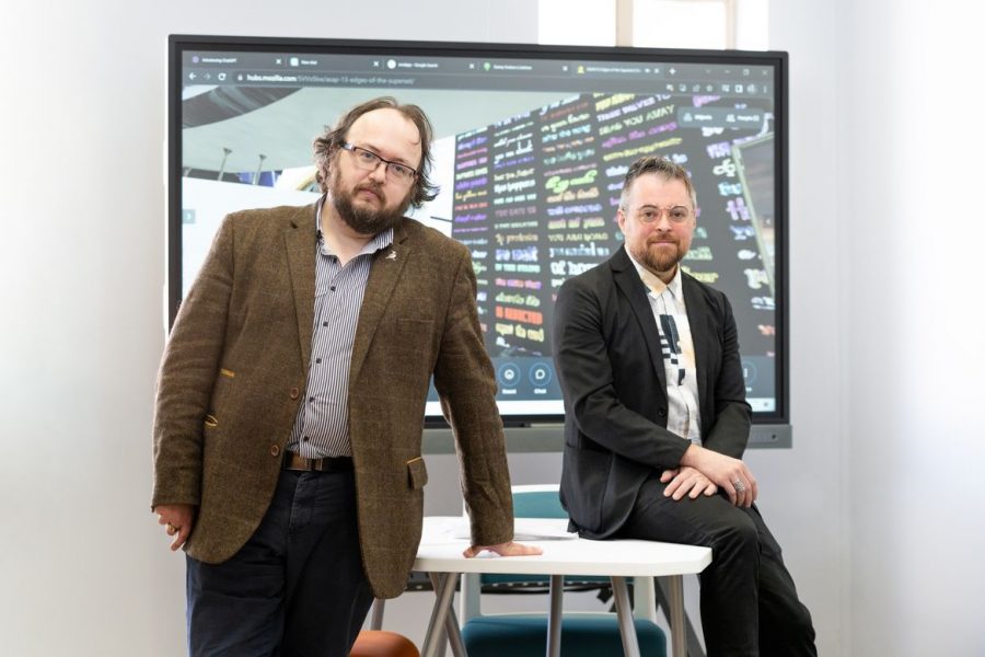 In front of a screen projecting lines of code, Jacob Foster leans against a table, while Danny Snelson sits on it.