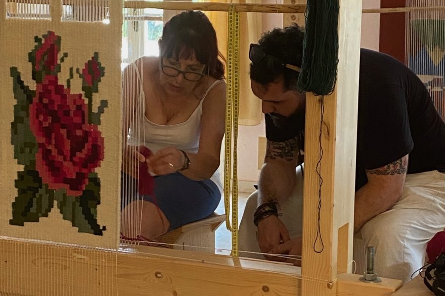 Two people bend over a loom. They are weaving an intricate red rose design.