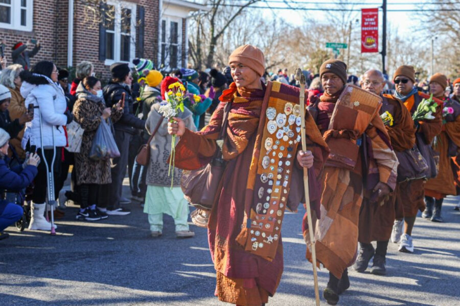 Buddhist monks in tan robes walk down a street as bundled-up supporters line sidewalk