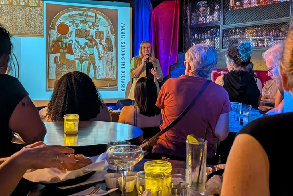 Marissa Stevens with microphone on stage at a bar, with backs of audience members' heads in foreground